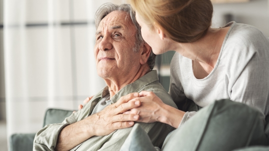 Adult woman offering support to an older man while sitting together on a couch indoors. | Adler Law